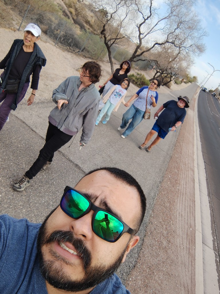 A group of six individuals walking together along a pathway lined with sparse vegetation and trees, with one person taking a selfie in the foreground.