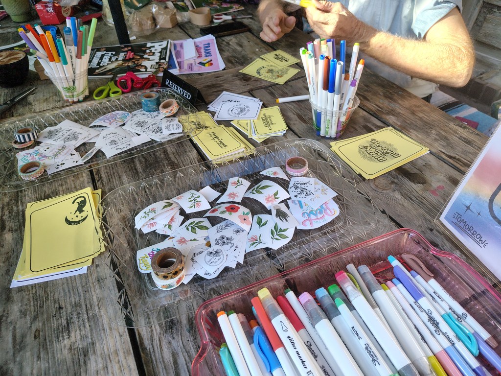 A table covered with various art supplies, including colorful markers, stickers, and washi tape, with a person's hands visible working on a project.