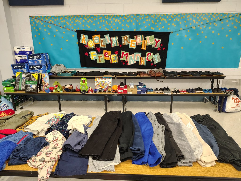 Tables filled with assorted clothing items, toys, and household goods for the Really Really Free Market event, with a colorful banner in the background.