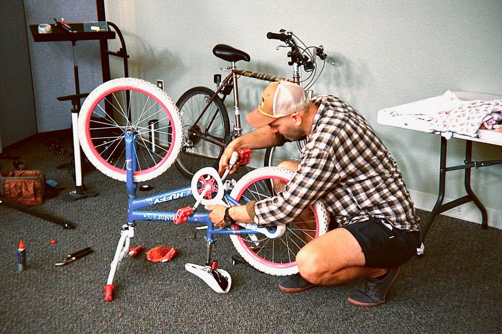 A person wearing a plaid shirt and hat is repairing a blue bicycle with pink wheels in an indoor space. Tools and bicycle parts are scattered around.