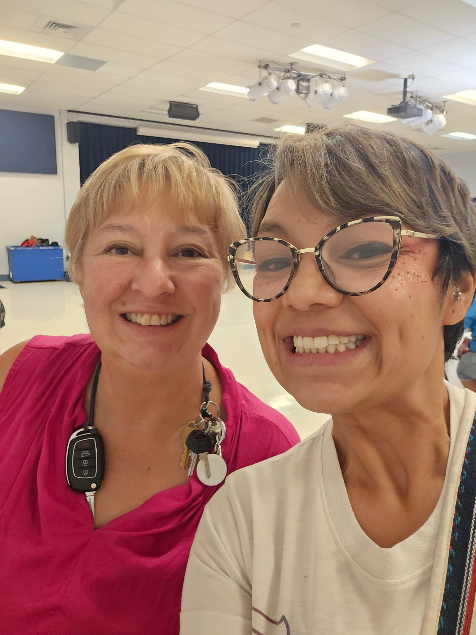 Two smiling women take a selfie in a brightly lit room. One wears glasses and a white shirt, the other a pink top with a key lanyard. The background shows ceiling lights and a blue wall.