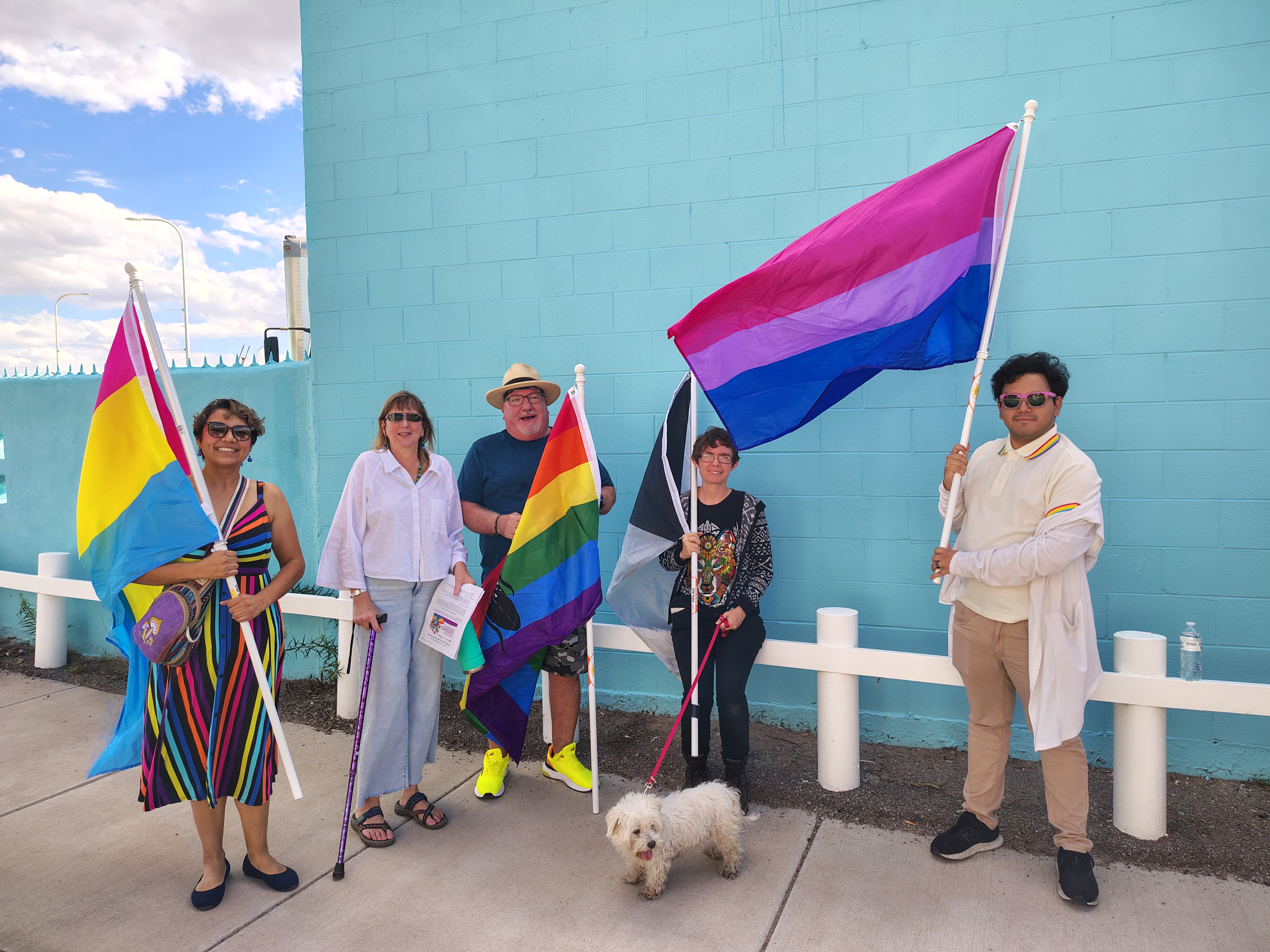 Five people standing in front of a blue wall hold various pride flags, including pansexual, rainbow, bisexual, and asexual. They appear joyful, with a small white dog in front.