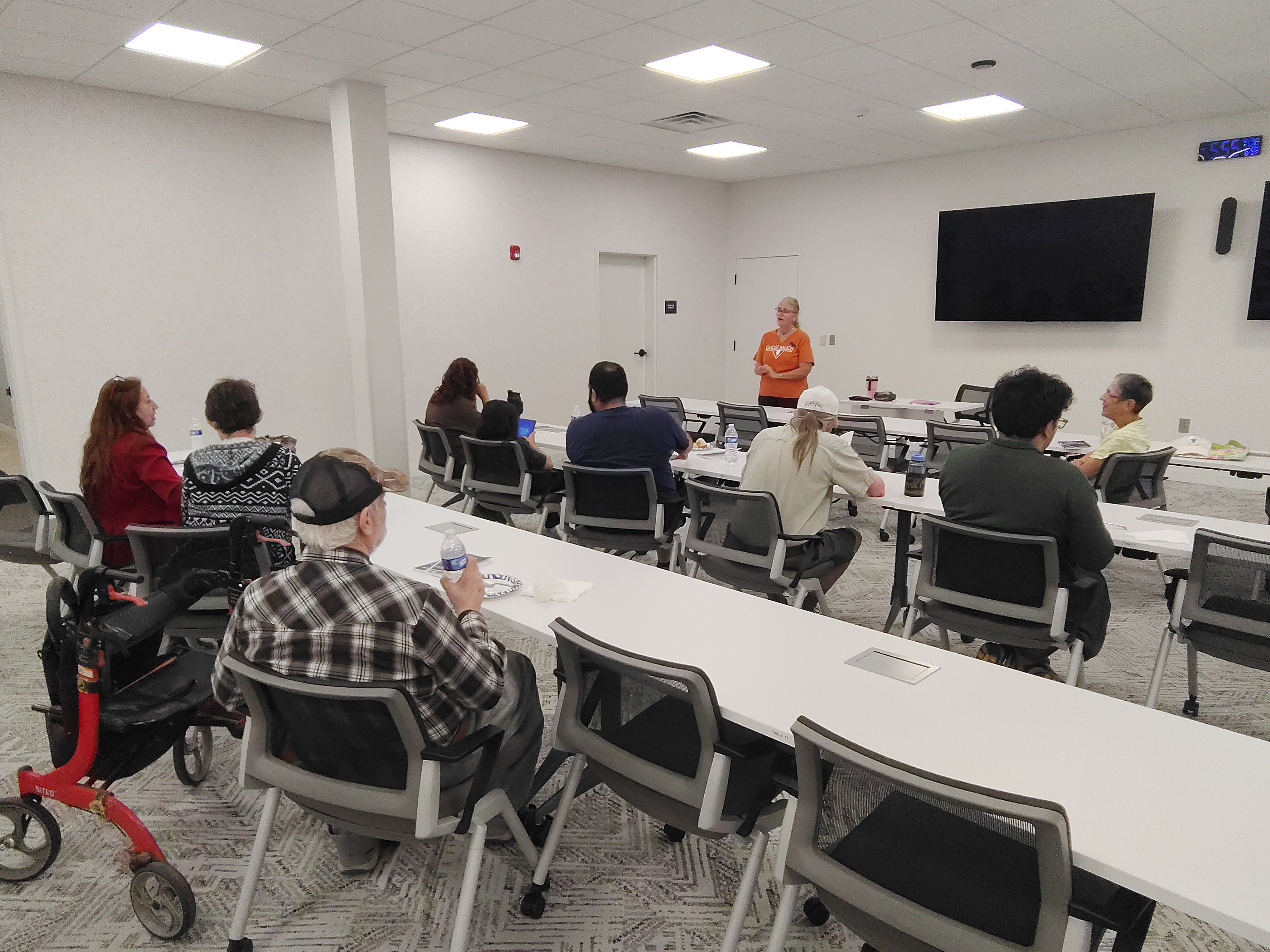 A group of people sit in a classroom with a woman standing at the front speaking. Chairs, a red walker, and a large screen are visible.