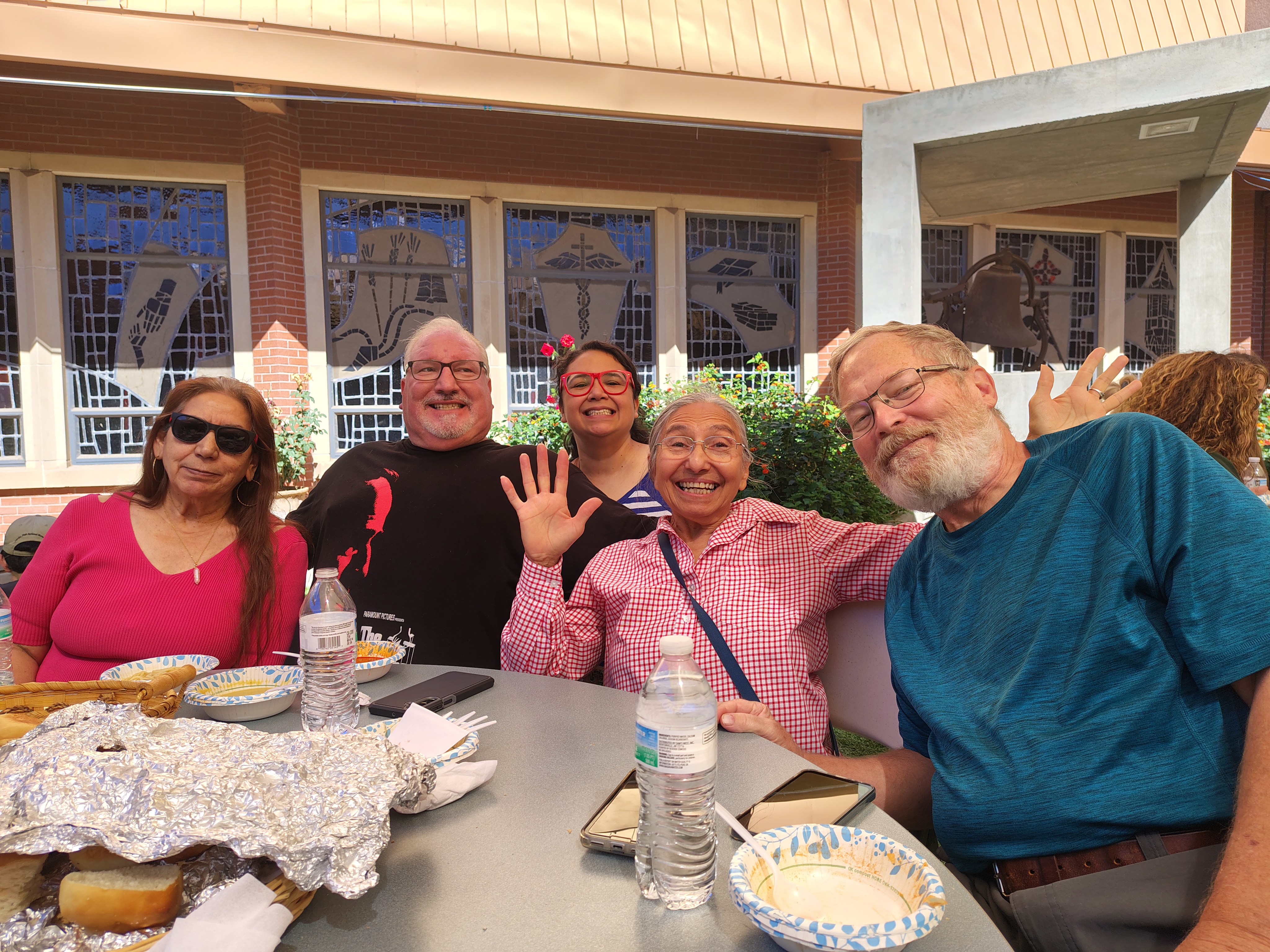A joyful group of five people sit at an outdoor table with bright sunlight, smiling and waving. The table has wrapped food items and water bottles.