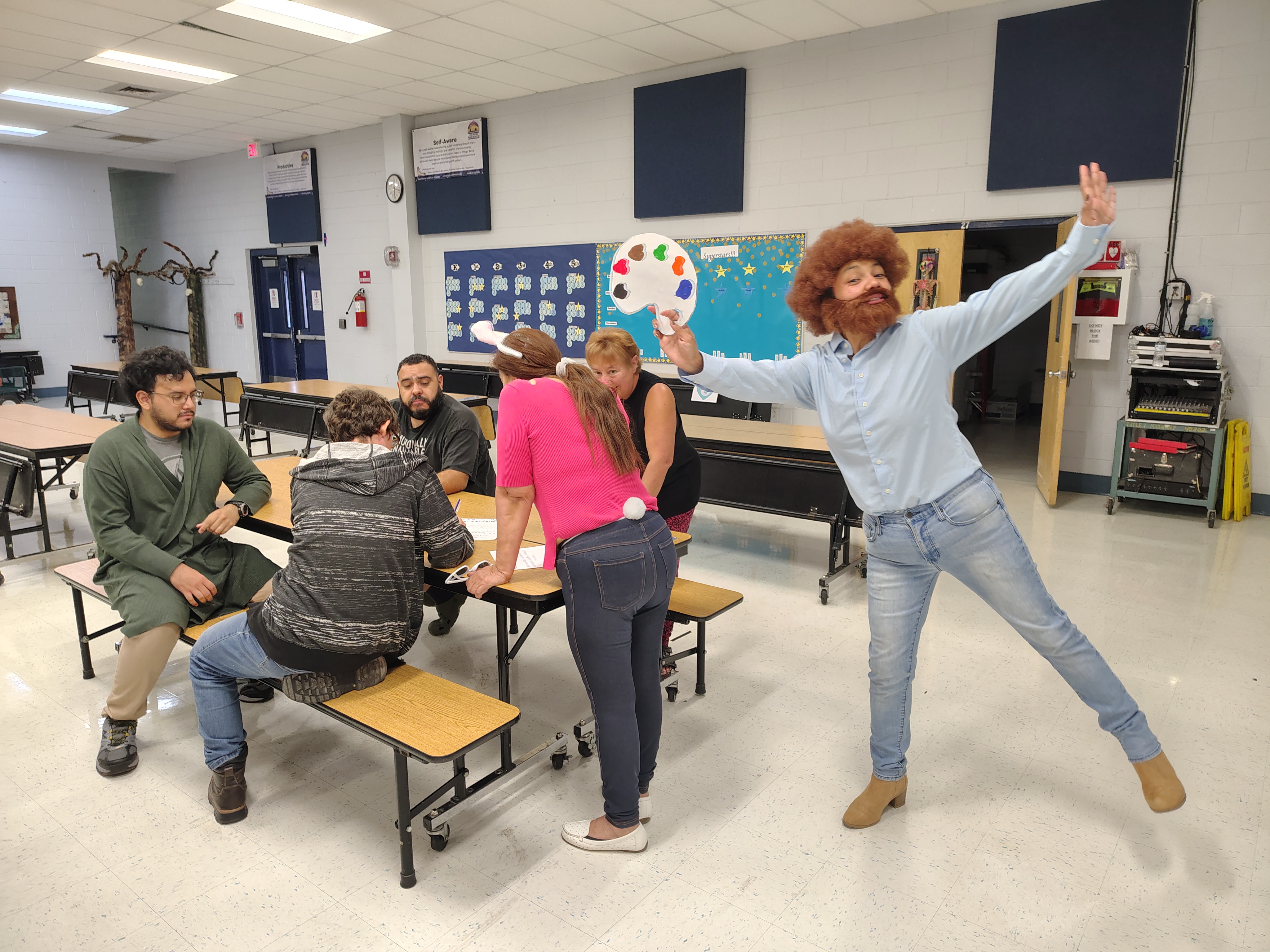 A group of people in costumes converse around a cafeteria table. One person, dressed as a painter with a wig and palette, poses playfully beside them. The atmosphere is lively and humorous.