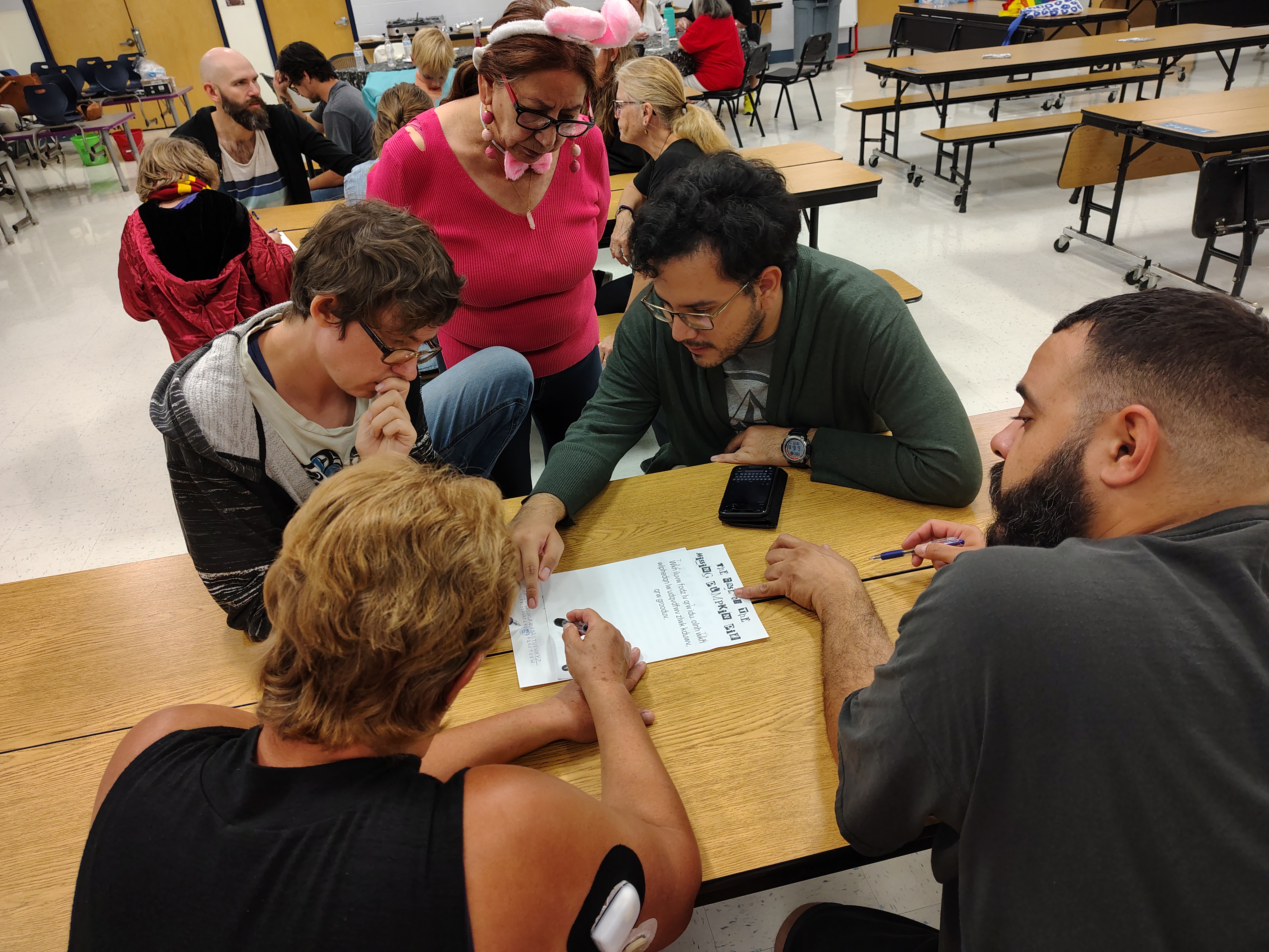 A group of five people huddle around a table in a classroom, focused on solving a sheet with text. One person wears pink bunny ears, suggesting a playful atmosphere.