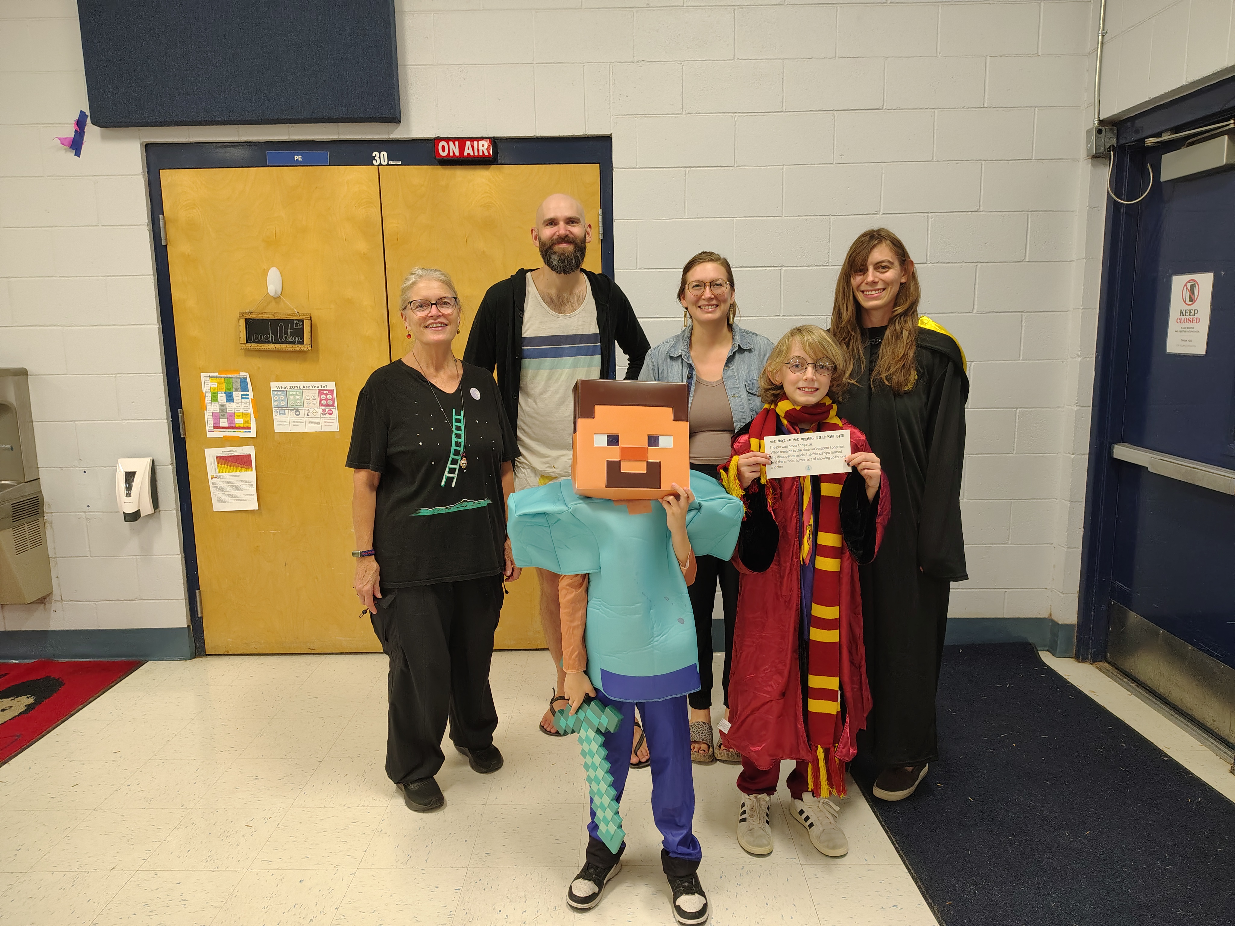 Group photo in a school hallway. Two children in costume, one as a Minecraft character and another in a wizard robe, stand with four smiling adults.