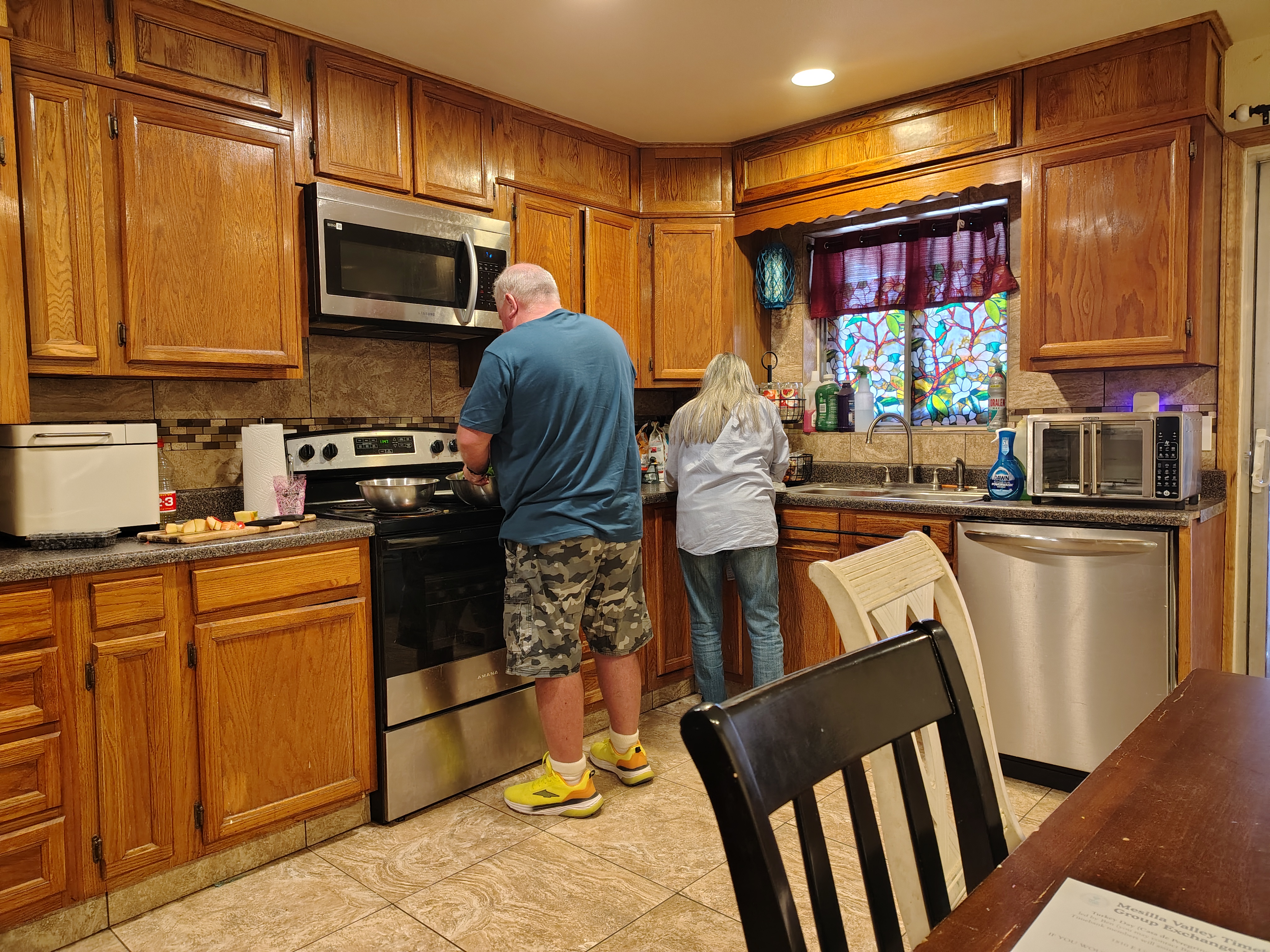 A man and woman cooking in a cozy kitchen with wooden cabinets. The man is at the stove; the woman is at the sink. Stained glass window adds color.