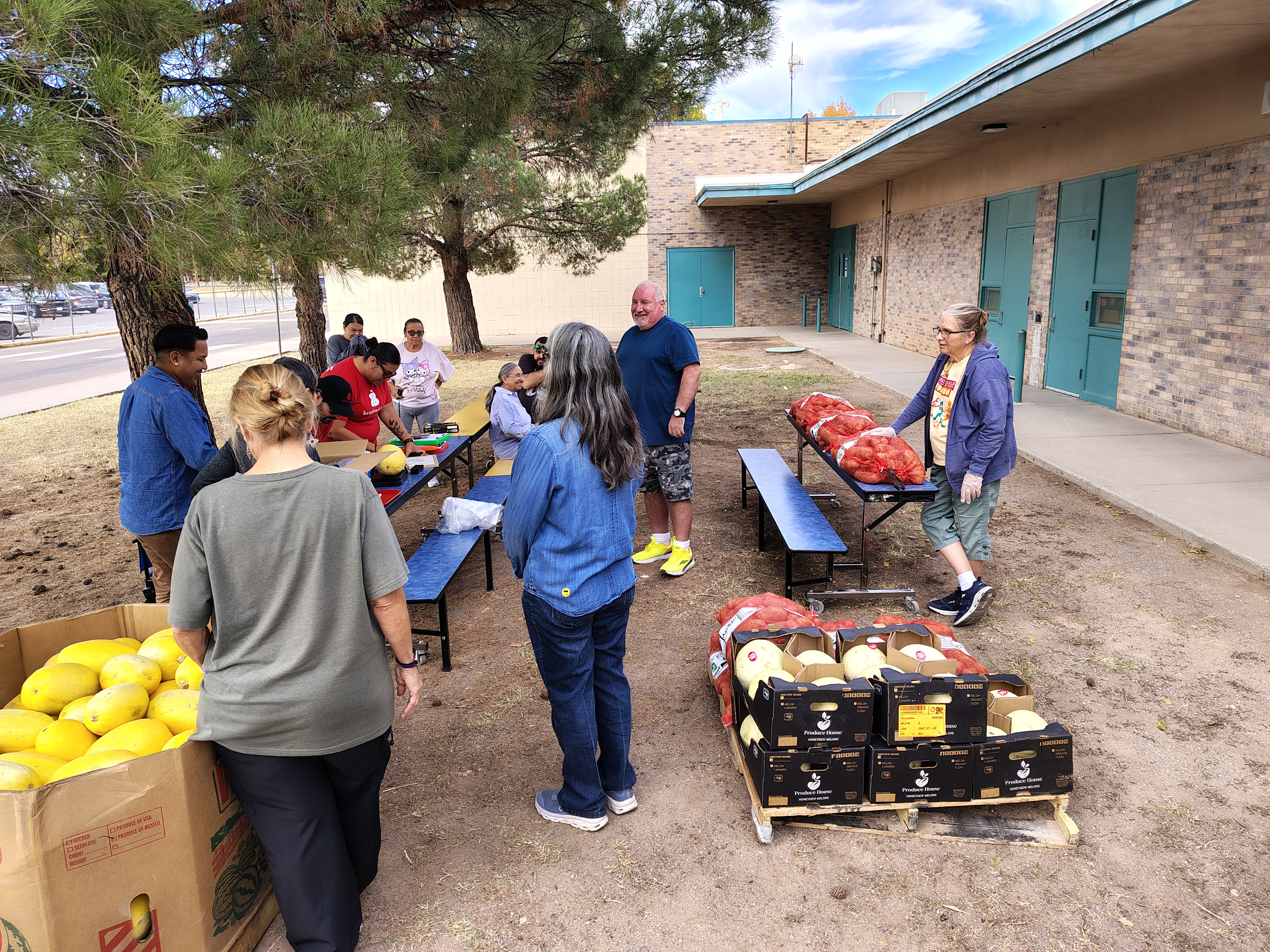 A group of people gather at outdoor tables near a brick building with teal doors, sorting fruit and vegetables. Trees provide shade, conveying a collaborative and community-focused atmosphere.