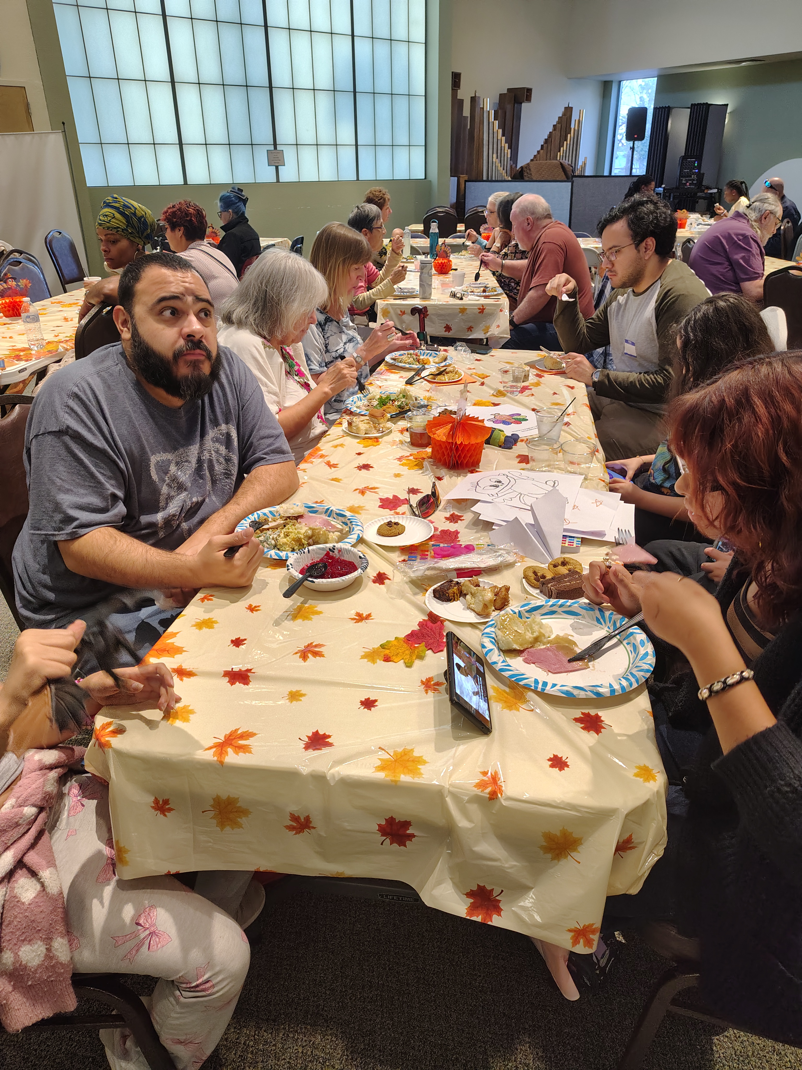 A diverse group of people, seated around a long table with autumn-themed decor, enjoy a meal together. The atmosphere is warm and communal.