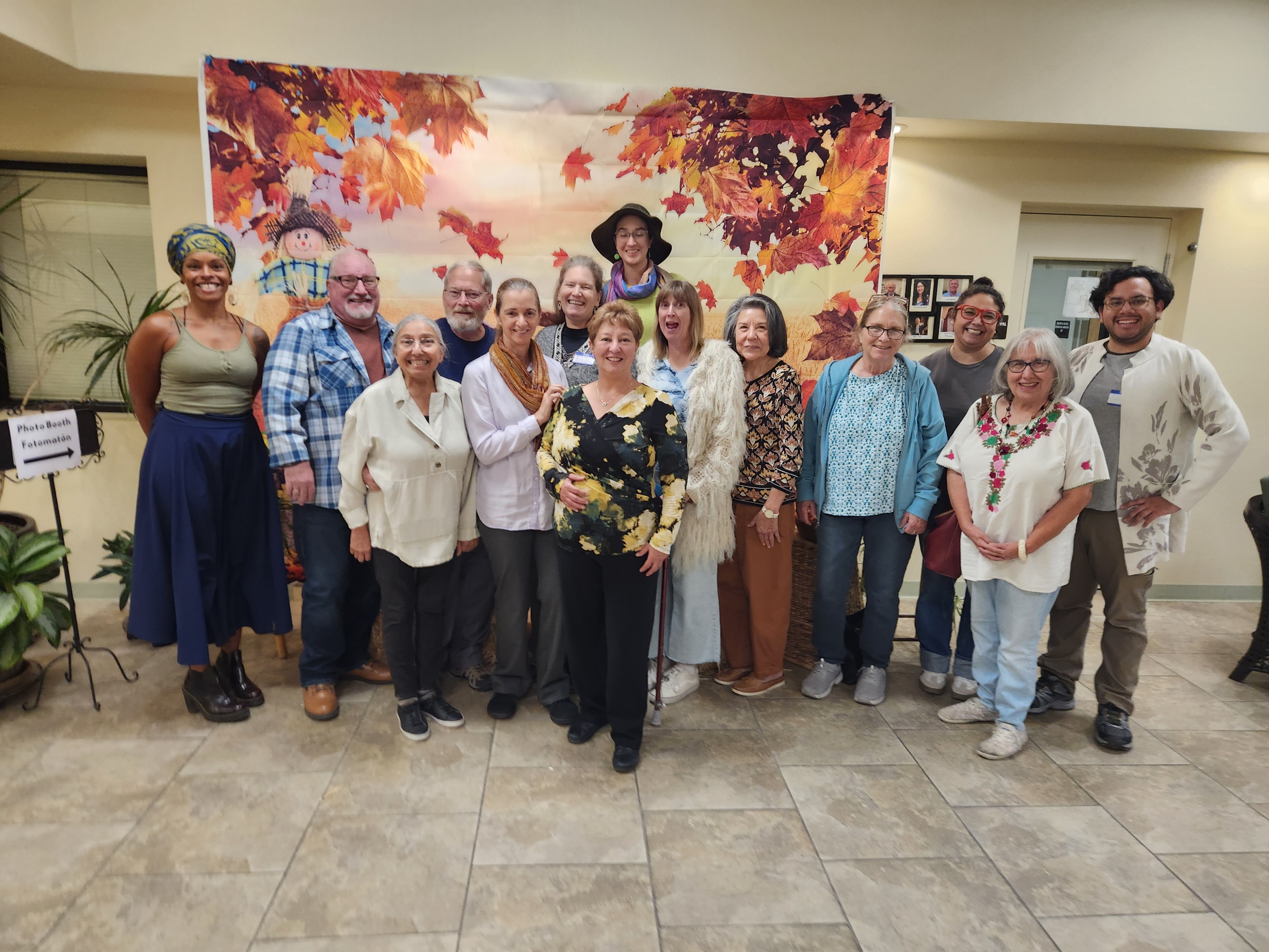 A group of fourteen people, smiling, pose in front of an autumn-themed backdrop with red and orange leaves. They are casually dressed, conveying a joyful and warm atmosphere.